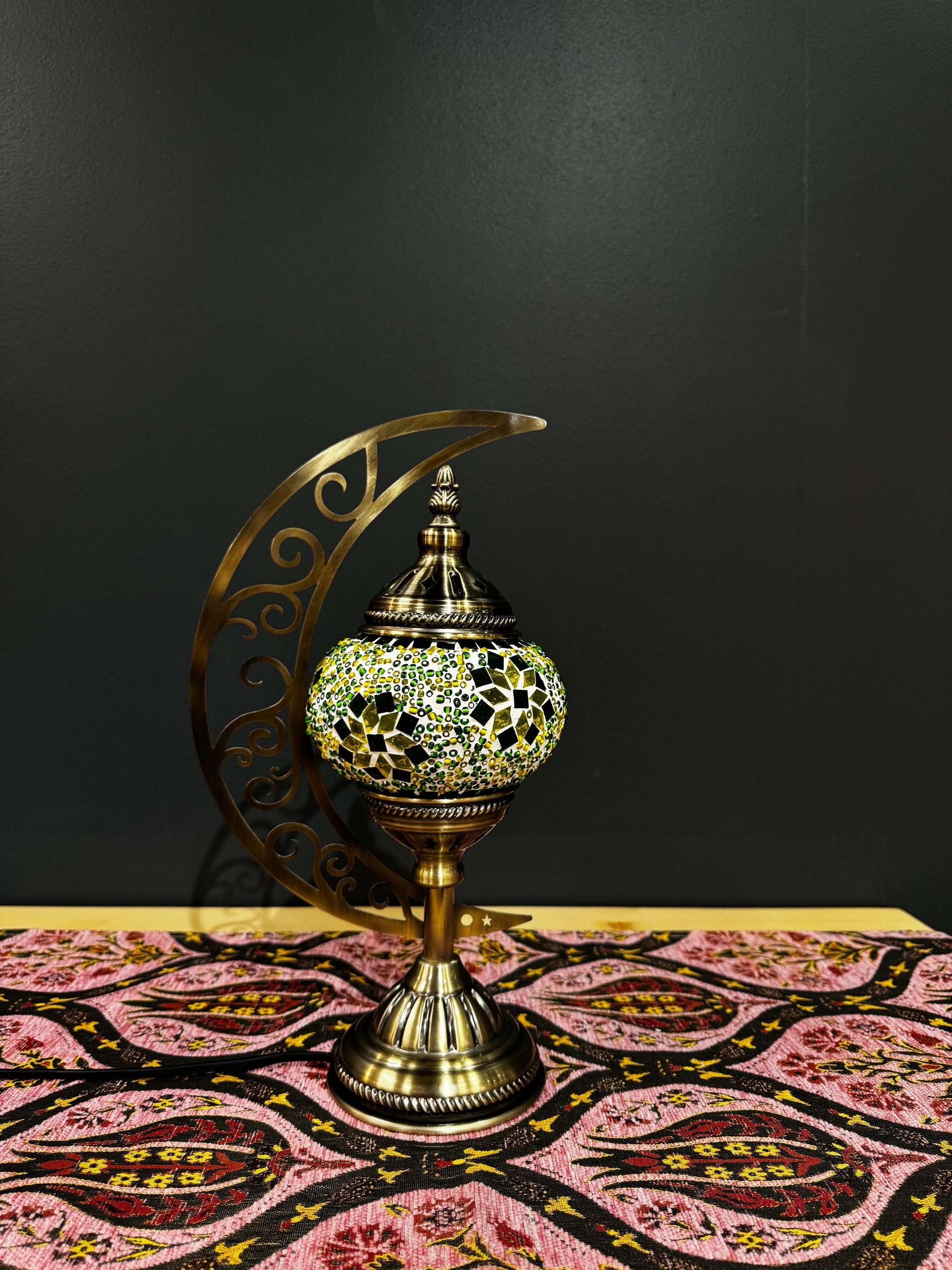 Emerald Glow Turkish Mosaic Lamp in crescent moon design with vibrant green and yellow mosaic on a patterned tablecloth.