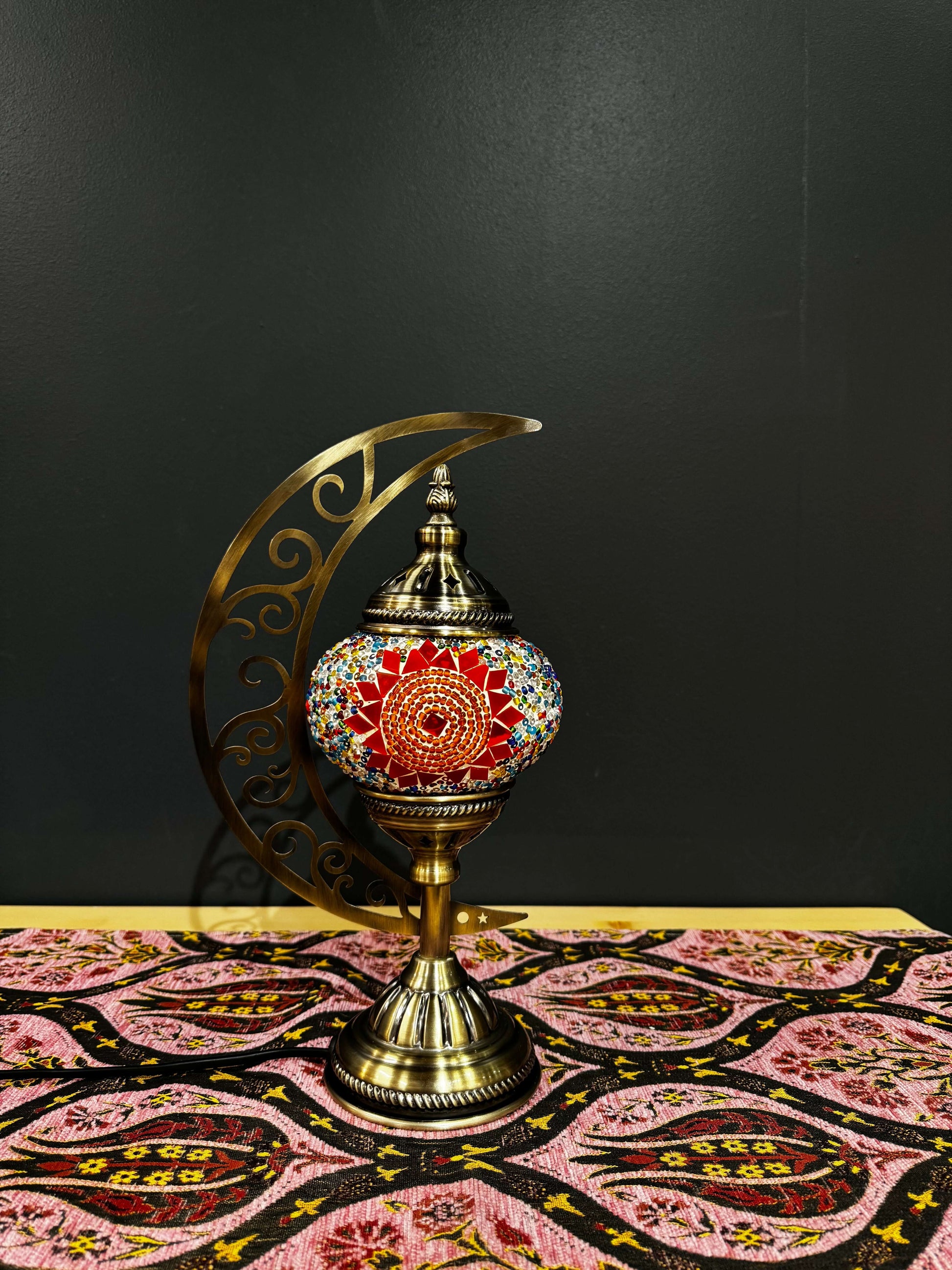 Sunburst Bloom Turkish Mosaic Lamp on a patterned tablecloth, featuring a crescent moon design and vibrant red mosaic.