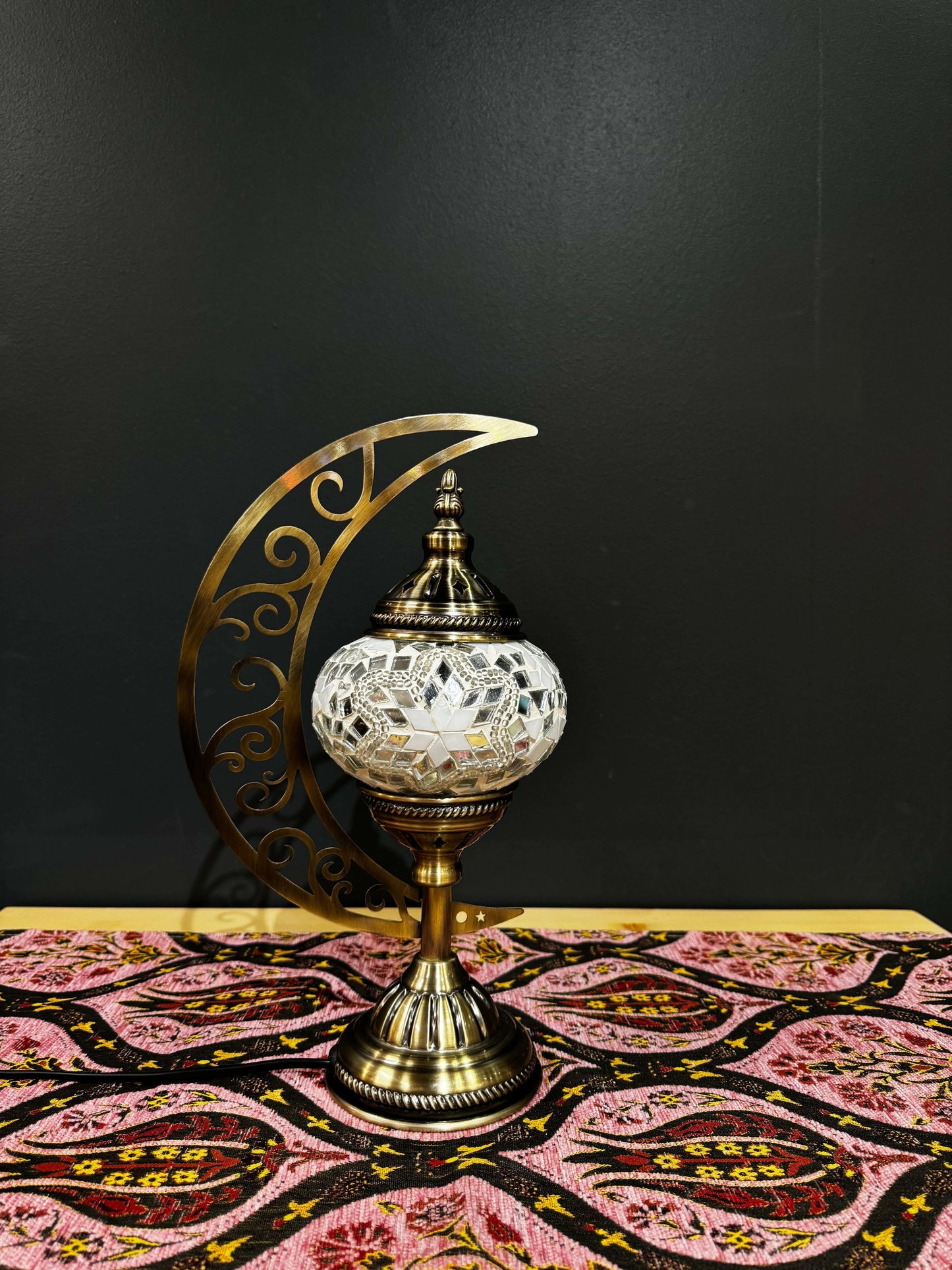 White Radiance Turkish Mosaic Lamp with crescent moon stand on colorful patterned tablecloth. Elegant home decor piece.