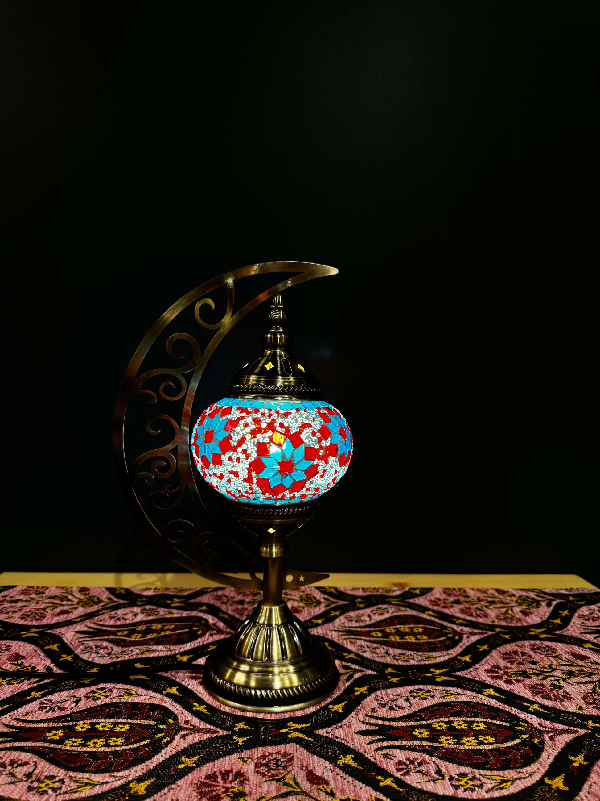 Colorful Ruby Blossom Turkish Mosaic Table Lamp with crescent moon stand, shining on an ornate patterned tablecloth.