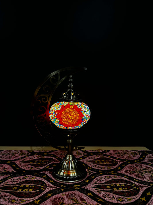 Sunburst Bloom Turkish Mosaic Lamp with crescent moon stand illuminating a vibrant floral design on a patterned tablecloth.