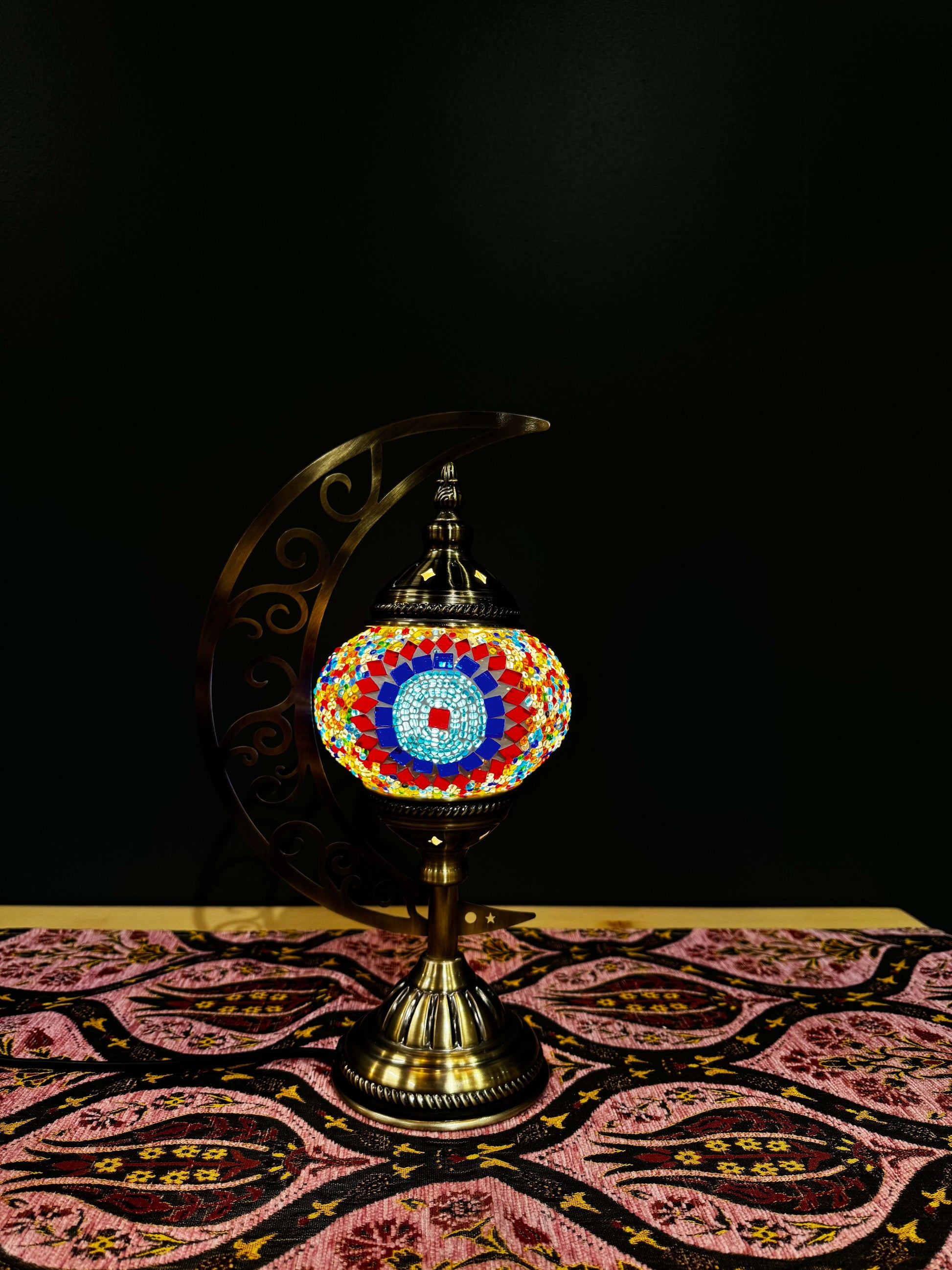 Sunburst Circle Turkish Mosaic Lamp with vibrant colors on a crescent moon stand, displayed on a patterned tablecloth.