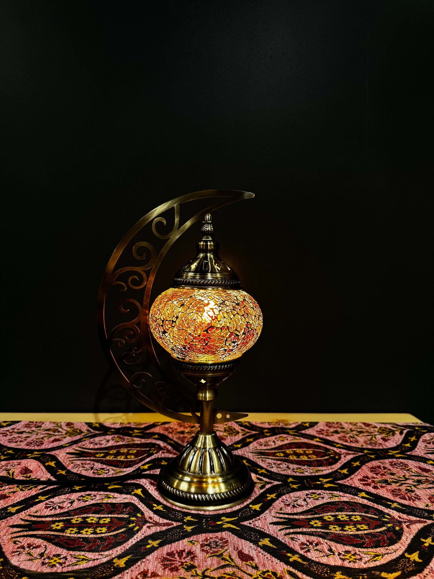 Amber Twilight Turkish Mosaic Lamp with crescent moon stand on patterned tablecloth, showcasing intricate amber mosaic design.